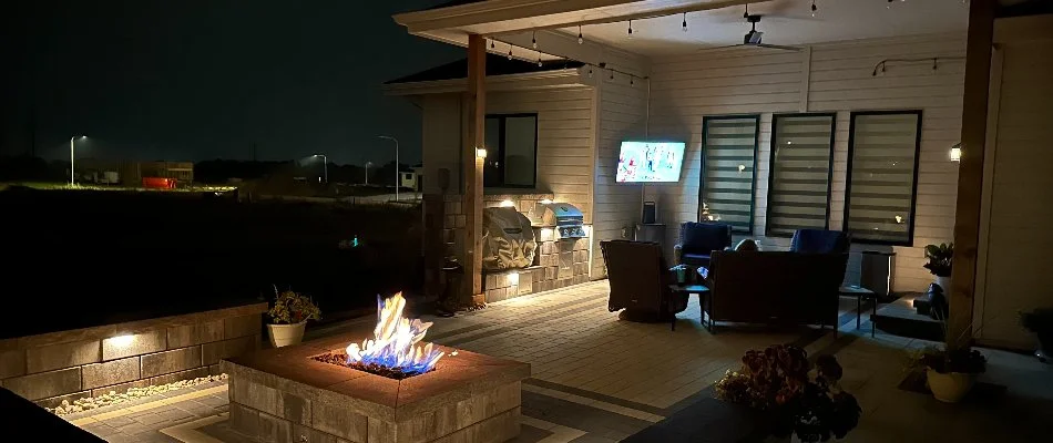 An outdoor kitchen in Omaha, NE, at night with outdoor lighting, a television, and a fire pit.